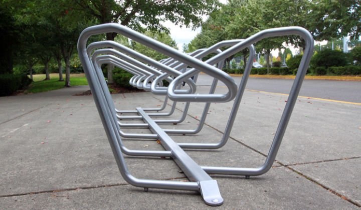 Low-angle view down a row of brushed steel Plaza bike rack loops on a sidewalk, trees behind