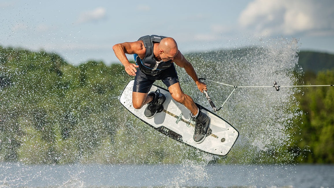 Wakeboarder in a life vest mid-air on a white Hyperlite board, spray flying behind him over a tree-lined lake